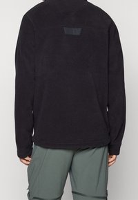 Giacca in pile nera con maniche lunghe, texture liscia e una piccola patch nera sulla parte superiore della schiena. Abbinata a pantaloni corti sportivi verdi.
