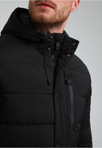 Czarna kurtka puchowa z zamykaną na zamek kieszenią na piersi, kapturem z regulacją i zapięciem na zatrzaski. Posiada pikowane sekcje i gładką fakturę materiału.