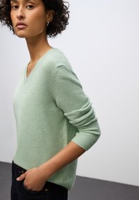 Světle zelený svetr s výstřihem do V z měkké pleteniny. Má dlouhé rukávy s žebrovanými manžetami a volný střih. Kombinovaný s tmavými džíny.
