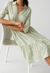 Robe chemise verte claire avec un col, motif blanc, longueur genoux avec une taille froncée et des manches trois-quarts, assise sur une chaise.