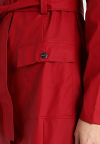 Czerwony płaszcz przeciwdeszczowy z paskiem w talii, dużą zapinaną na guziki kieszenią i gładką, wodoodporną teksturą.