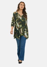 Vrouw in een asymmetrische tuniek met groen, bruin en witte patronen, met driekwart mouwen, blauwe jeans en zwarte flats, die vooruit loopt.