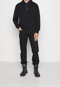 Sort pullover med lynlåsfrynse, kombineret med sorte bukser og snøreløse sorte støvler med metaløjer og teksturerede såler.