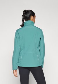 Jachetă softshell teal cu guler înalt, design ajustat, buzunare laterale cu fermoar și detalii de cusătură albă, purtată peste o pereche de pantaloni negri.