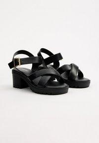 Sandalias de tacón negro con un diseño de tira ancha, fabricadas en cuero suave. Los zapatos tienen una suela gruesa y un acento de hebilla metálica.