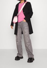 Schwarzer Mantel, rosa Strickpullover, graue Cargohose mit weißen Spritzern und klobigen schwarzen Plateauschuhen. Kleine rosa Tasche mit Schulterriemen.
