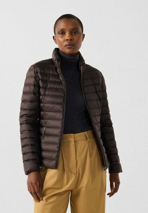 Chaqueta acolchada marrón con cuello alto, cremallera frontal y mangas largas, combinada con un jersey de cuello alto oscuro y pantalones beige.
