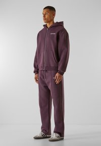 Veste à capuche zippée violette avec poches avant, assortie à un pantalon de survêtement avec cordon de serrage. Les détails incluent une couture légère et un logo.
