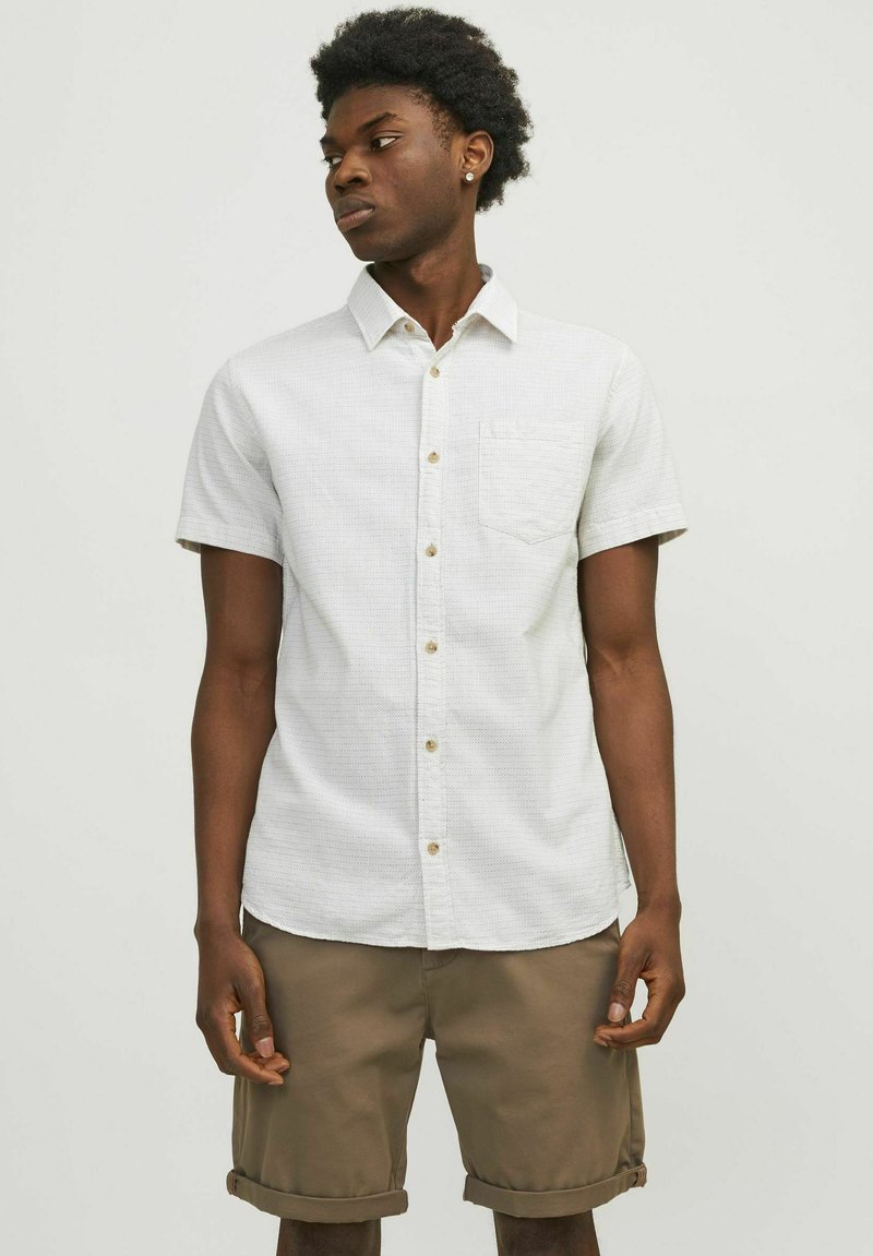 Camisa de botón de manga corta de color claro con un acabado texturizado y un bolsillo en el pecho, combinada con pantalones cortos beige remangados.
