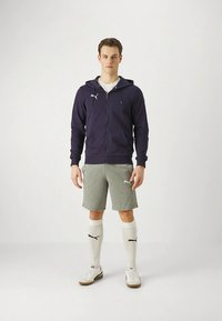 Marineblå hettegenser med glidelås, grå shorts, hvite sokker med svarte detaljer og hvite joggesko. Hettegenseren har en logo på brystet; shortsen har en logo på siden.