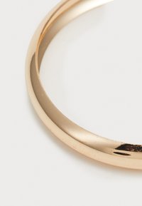 Kuldse tooniga metallist bangle, millel on sile ja poleeritud pind. Ümara kujuga, kergelt kaarduvad servad, esitlevad minimalistlikku disaini ja peeneid kriime.