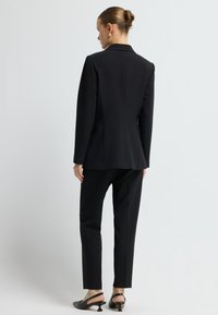 Czarny garnitur z dopasowaną marynarką i prostymi spodniami. Posiada wycięty kołnierz i gładką tkaninę. Uzupełniony czarnymi klapkami na obcasie.