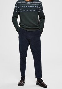 Mörkgrön tröja med ett vitt geometriskt mönster vid axeln, ihop med mörkblå kordbyxor och blanka svarta finskor.