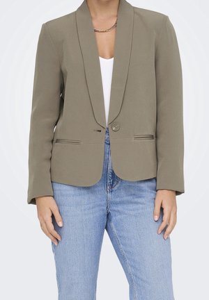 Person wearing a taupe single-button blazer over white top and light blue jeans, with a gold chain necklace visible at the neck.