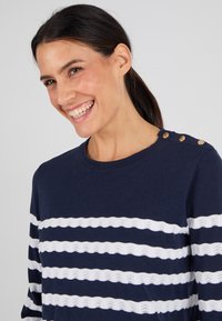 Maglione blu navy con strisce orizzontali a motivo onda bianche. Presenta un collo rotondo e dettagli con bottoni dorati sulla spalla. Texture morbida.