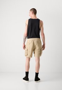 Canotta nera da uomo abbinata a pantaloni cargo beige, con tasche frontali, tessuto testurizzato e vita elastica; scarpe e calzini neri.