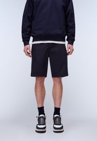 Man draagt zwarte korte broek, zwarte sokken, zwart-witte sneakers, marineblauwe trui en grijs ondershirt, staand tegen een lichte achtergrond.