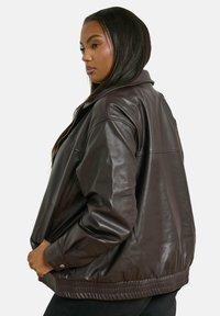 Giubbotto bomber in pelle marrone con vestibilità rilassata, vita a coste, tasche laterali e colletto. Texture liscia e design minimalista.