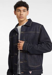 Dark blue denim jacket with a button-up front, chest pockets, and contrast stitching. Smooth texture, casual design, and logo on the hem.