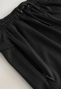 Shorts de sport noirs en tissu texturé, avec une taille élastique dotée d'un cordon de serrage et des poches latérales zippées.