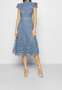 Sukienka z koronką w przykurzonym niebieskim kolorze, z krótkimi falbaniastymi rękawami, dopasowanym gorsetem i rozszerzanym dołem. Charakteryzuje się złożonymi wzorami florystycznymi i wysokim dekoltem.