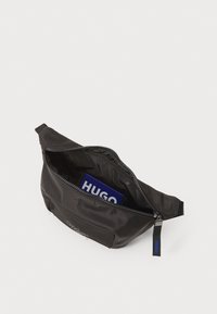 Borsa in tessuto nera con chiusura a zip, con un logo sul fronte e un oggetto blu parzialmente visibile all'interno. Texture liscia.