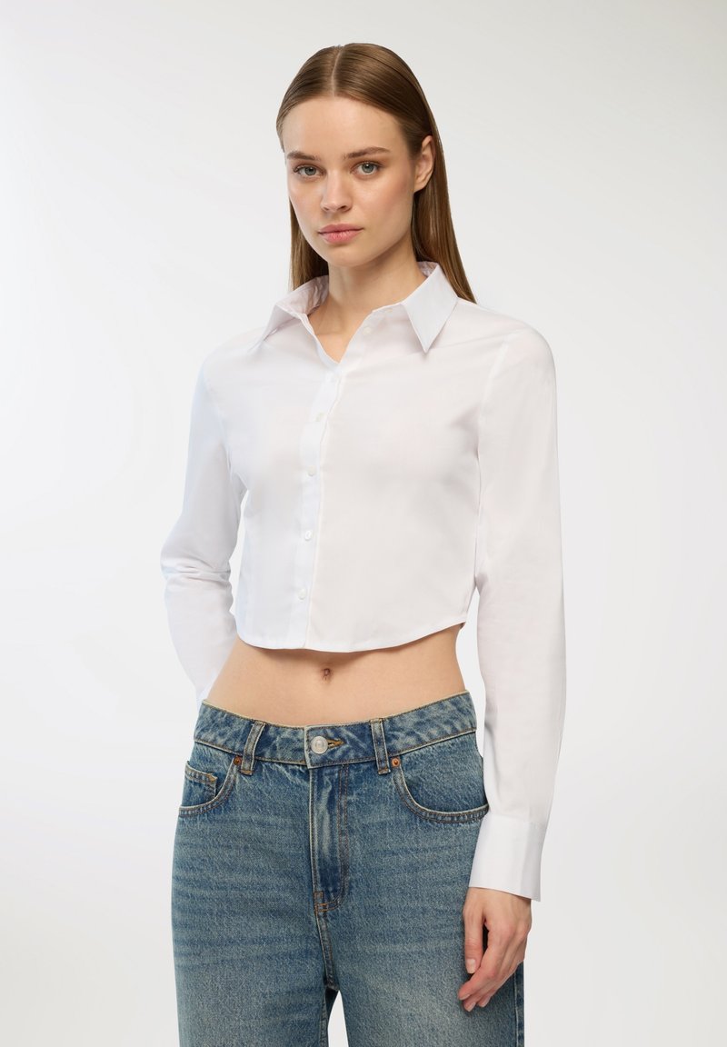 Camicia bianca cropped con maniche lunghe, realizzata in tessuto liscio; presenta un colletto classico e un orlo dritto. Indossata con jeans in denim blu.