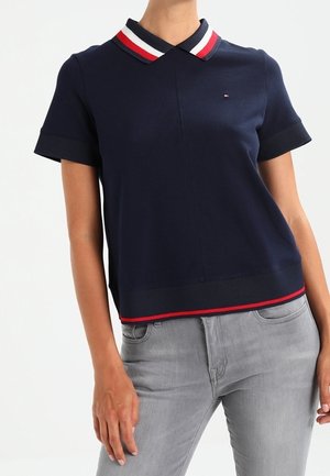 Granatowa polo z krótkim rękawem, z czerwono-biało-niebieskim paskowanym kołnierzem i wykończeniem, noszona z jasnoszarymi dżinsami.