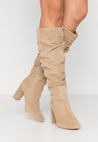 Botas de ante beige hasta la rodilla con un diseño holgado, punta afilada y tacón cuadrado. Presenta una cremallera lateral para facilitar su uso.