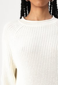 Pull en maille blanche avec un motif texturé, présentant un col rond et des manches longues, mettant en valeur un col côtelé et un design raglan.