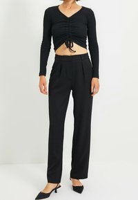 Czarny długi rękaw crop top z marszczonym przodem i detalem w postaci wiązania, zestawiony z czarnymi spodniami o wysokim stanie z zakładkami i zwężanymi nogawkami, noszonymi z czarnymi butami na obcasie.