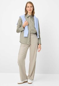 Gestreept olijfgroen overhemd, lichtblauwe trui over de schouders gedrapeerd, beige wijde broek met streep aan de zijkant, witte sneakers. Eenvoudige, op maat gemaakte look.