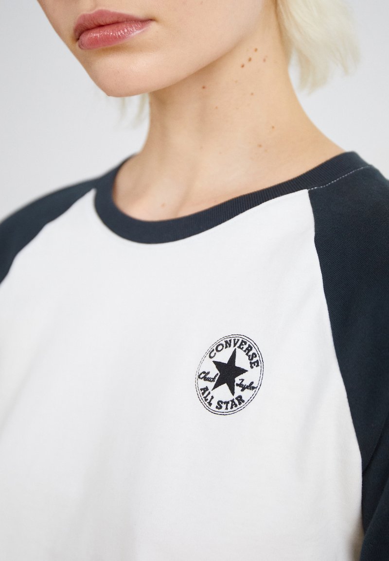 T-shirt bianco con maniche raglan blu scuro e un logo nero rotondo con una stella e la scritta "Converse All Star" ricamato sul petto.
