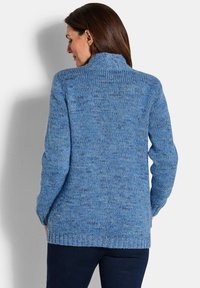 Frau, die einen blauen gestrickten Pullover mit langen Ärmeln und hohem Kragen trägt, steht mit dem Rücken zur Kamera.