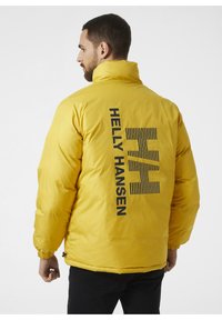 Żółta kurtka puchowa z wysokim kołnierzem, z czarnym logo "Helly Hansen" na plecach oraz poziomymi paskami akcentującymi.