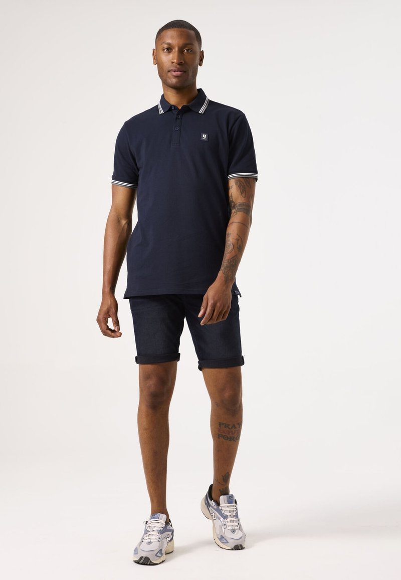Navy-Polohemd mit weiß gestreiftem Kragen, kombiniert mit dunklen Jeansshorts. Mit kleinem Logo auf der Brust und umgeschlagenen Shorts-Säumen.