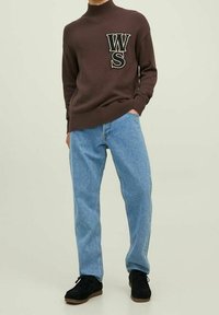 Bruine gebreide trui met ribbelkraag en grote "WS" letters op de borst. Gecombineerd met lichtblauwe recht-toe-recht-aan jeans en zwarte schoenen.