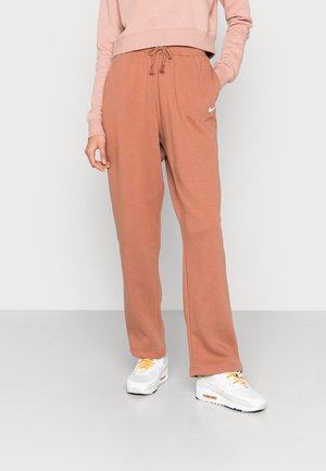 Person iført brune Nike træningsbukser, lyserød sweatshirt og hvide sneakers med orange snørebånd, der står på en ensartet gulv.