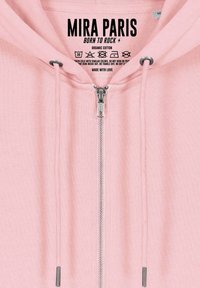 Felpa con zip di colore rosa chiaro realizzata in cotone biologico, con zip in metallo, coulisse con accenti metallici e istruzioni per la cura stampate all'interno.