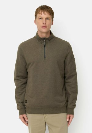 Man staat recht naar voren kijkend, gekleed in een olijfgroene trui met een kwart rits en beige broek tegen een effen witte achtergrond.
