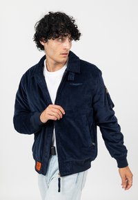 Marineblauwe corduroy jas met een rits, ribbels aan de mouwen en twee zijzakken; voorzien van een klein logo op de borst en een oranje label.