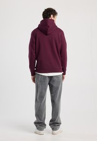 Bordeauxfarbener Kapuzenpullover mit weichem Material und gerippten Bündchen, getragen über einem weißen Hemd. Kombiniert mit locker sitzenden grauen Jeans und weißen Turnschuhen.