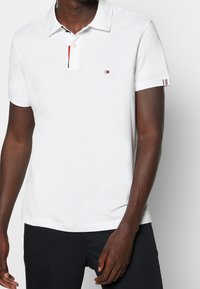 Polo shirt blanc en coton avec une patte de boutonnage à deux boutons. Présente un accent coloré au niveau du col et des accents rayés sur les manches.