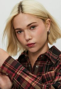 Plaid flannel shirt featuring red, black, and tan colors, with snap buttons and a classic collar. Short, straight blonde hair.