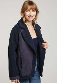 Veste bleu marine à double texture avec un extérieur doux et shaggy, un intérieur lisse, un col à revers et une poche zippée sur le côté.