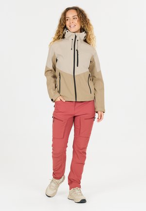 Frau mit lockigem Haar, die eine beige und braune Jacke mit mehreren Reißverschlusstaschen sowie rote Cargo-Hosen und beige Wanderschuhe trägt.