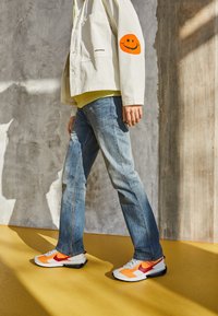 Baskets blanches avec des accents orange et rouge, associées à un jean bleu et une veste blanche ornée d'un patch visage souriant orange.