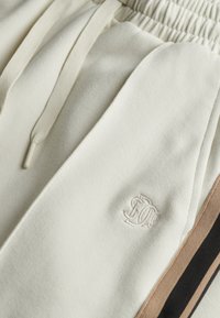 Witte katoenen sweatpants met een elastische tailleband, trekkoord en geborduurd logo. Accenten zijn bruin en zwarte zijstrepen.