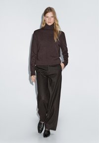 Donkerbruine gebreide coltrui gecombineerd met zwarte wijde broek en zwarte schoenen; gladde textuur met een minimalistisch ontwerp.