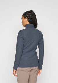 Vrouw met gevlochten haar, gekleed in een getextureerde donkergrijze langmouwtop en beige broek, met haar rug naar de camera, tegen een effen lichte achtergrond.
